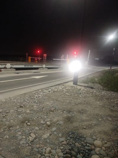 The image captures a nighttime scene at a checkpoint on the Түндүк айланып өтүүчү жол 84 highway in Kyrgyzstan. A traffic light displays a red signal, indicating a stop. A barrier gate, likely part of the checkpoint infrastructure, is visible. In the foreground, the road surface is marked with a white arrow pointing to the right, suggesting a lane designation. The ground is covered with various sizes of gravel and stones. A bright light source, possibly from a vehicle or streetlamp, illuminates the area. A sign on a long, horizontal structure reads "METTLER TOLEDO," identifying a company. The background is dark, with hints of distant lights and vegetation.