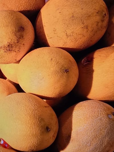 The image is a close-up, overhead view of a pile of cantaloupes. The fruits are predominantly a light orange color with a netted rind texture. The lighting creates strong shadows, highlighting the round shapes of the melons and the texture of their skins. There are no people or discernible background elements indicating a specific city or location.