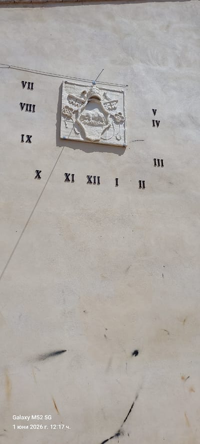 The image shows a sundial mounted on a wall. The sundial is a stone relief with an ornately decorated face. Roman numerals are etched into the wall around the sundial, marking the hours. A metal pointer extends from the sundial, casting a shadow that indicates the time. The wall has a rough, textured surface and is painted in a light beige or cream color. There are some dark stains and marks on the wall. The bottom right corner of the image contains a timestamp indicating that the photo was taken on "1 юни 2026 г. 12:17 ч." with a Galaxy M52 5G phone. Based on the subject, the sundial, the location might be Spain, Canals.