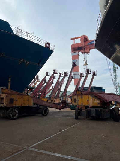 The image depicts a shipyard in Geoje-si, South Korea, with the hull of a large ship dominating the left side of the frame. In the foreground, several yellow and red boom lifts are parked in a line, their articulated arms extended upwards. These lifts appear to be industrial equipment used for construction or maintenance. Behind the lifts, a massive orange gantry crane, branded with "SAMSUNG", looms over the scene. The crane is positioned above the ship's hull. The sky is overcast with a bright, diffused light. The ground is paved with concrete, showing some wear and tear. The overall impression is one of a large-scale industrial operation in progress.
