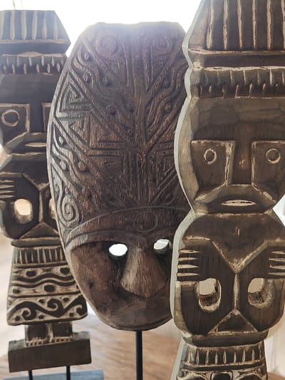 The image shows a close-up, vertically oriented view of several intricately carved wooden masks. The masks appear to be ancient or tribal in origin, with one prominently displayed in the foreground. This central mask is oval-shaped and features detailed geometric patterns and stylized facial features, including two eye openings. The wood has a rich, dark brown patina, and some areas are highlighted with a lighter, possibly white or light gray, accent color, suggesting age or a deliberate decorative technique.
Behind the central mask, parts of other masks are visible, each with distinct carving styles. One mask above the central one has a more abstract, angular design with circular and elongated openings for eyes. Another mask below the central one exhibits a different pattern of parallel lines and geometric shapes. The texture of the wood is evident, showing the marks of carving tools.
The lighting in the image is strong and suggests it might be daytime, possibly outdoors or in a well-lit indoor space. The bright light causes some areas of the masks to be overexposed, particularly on the left side, and creates highlights and shadows that emphasize the depth and detail of the carvings.
The overall impression is one of artistry and cultural heritage. The masks are arranged in a way that suggests they are either part of a collection, displayed for exhibition, or perhaps part of a larger decorative installation. There is no visible text within the image. The setting appears to be in Maitland, United States of America, but specific location cues within the image are absent.