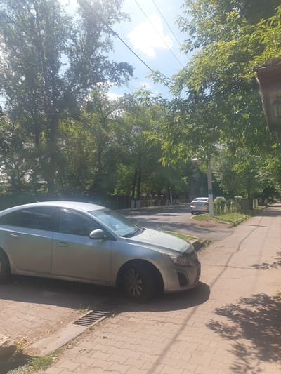 A silver sedan is parked on a brick sidewalk next to a street in Petropavl, Kazakhstan. The car is facing right, with its front on the right side of the frame. The street is lined with trees and a sidewalk. In the background, there are other cars and buildings. The sky is blue with some clouds. The lighting suggests it is daytime.
