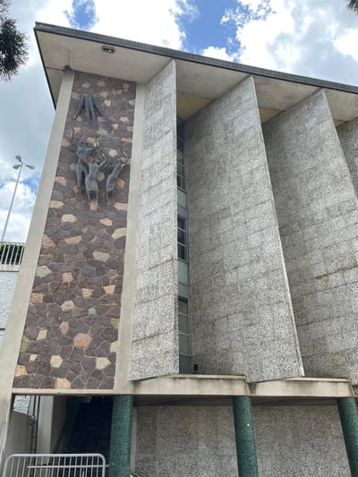 The image presents a low-angle, upward view of a building's facade, likely in Brisbane City, Australia, showcasing its architectural details. The primary features are several prominent horizontal concrete slabs, possibly balconies or sunshades, textured with a speckled, light-colored aggregate finish. These slabs are stacked, creating strong parallel lines and a sense of depth. A strip of windows is visible between two of the lower concrete tiers.

Below the lowest concrete slab, a section of the building's exterior wall is decorated with an irregular mosaic of multi-colored stone tiles, featuring shades of brown, beige, green, and grey. Mounted on this stone-clad wall is an abstract metal sculpture or relief, dark in color, depicting stylized forms that resemble a herd of animals, possibly cattle or antelopes, in motion.

To the right, a thick concrete column runs vertically, complemented by two cylindrical structures covered in green, possibly tiled, material. The upper left portion of the image reveals a blue sky with scattered white clouds, indicating a bright, clear daytime. Elements of a street light pole and a white railing are partially visible at the bottom, along with some dark green foliage. There is no visible text or human activity in the scene.