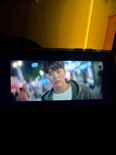 In a dimly lit environment, reportedly within the city of Lubnya, Russia, a dark-colored mobile device, oriented horizontally, displays video content. The device's screen exhibits multiple fine scratches and a noticeable diagonal crack across the upper right portion. The video features a young East Asian male, positioned slightly off-center to the right, looking forward with a wide-eyed, alert expression. He has short dark hair and is dressed in a white t-shirt beneath an open grey hooded jacket. The background within the video is an out-of-focus, brightly lit urban night scene, characterized by numerous bokeh light sources in various colors, including white, pink, blue, and orange, suggesting a crowded street or market. Indistinct, blurred human figures are visible in the background to the left. The immediate environment surrounding the mobile device is predominantly dark, with a distinct warm yellow-orange light illuminating the upper right background, reflecting off an unseen vertical surface.