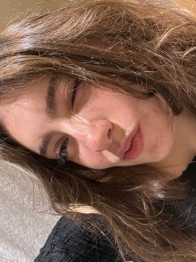 A close-up, selfie-style photograph shows a light-skinned person with light brown, wavy hair. The photograph is taken from a slightly above angle, with the person's head tilted. One eye is open, looking at the camera, while the other is squinting shut. A small gold ring is visible in the nostril. No specific location in Hull, United Kingdom can be identified from the visual information provided. The background is slightly blurred, showing a mix of the person's hair and a dark, textured surface.