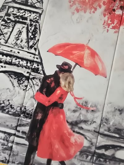 A painting depicts a couple embracing under a red umbrella. The man wears a black coat and hat, while the woman is in a red dress. Behind them, the Eiffel Tower is rendered in black and white. The scene is set against a backdrop of red leaves, suggesting an autumn setting. The artwork is divided into panels, with a visible seam running vertically down the center of the image. The location in Arshan, Russia is not ascertainable from the provided image.