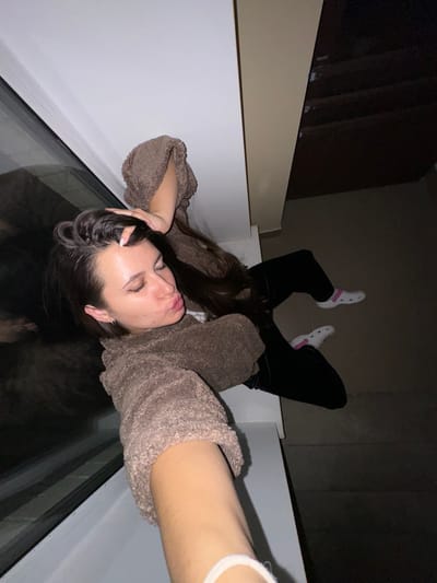 A young woman is lying on a glass surface, likely a skylight or similar architectural feature, within a building. She is wearing black pants and white socks with pink accents, and a brown, textured sweater. The surrounding environment appears to be an interior with beige walls and a dark, possibly wooden structure in the background. The perspective is angled, as if the photograph was taken from directly above, looking down through the glass. It cannot be determined the location in the city of Akulovo (Gp Kubinka), Russia.
