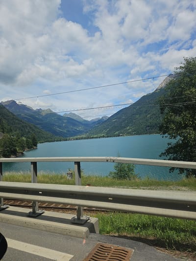 The image captures a scenic view of a turquoise lake nestled amidst lush green mountains under a partly cloudy sky. In the foreground, a metallic guardrail runs horizontally across the frame, separating a paved road from a grassy embankment and railway tracks. Beyond the guardrail, the expansive lake dominates the midground, its calm surface reflecting the surrounding landscape. The background is characterized by a range of imposing mountains, some with snow-capped peaks, densely covered with evergreen forests. The sky is a vibrant blue, dotted with fluffy white clouds. The location appears to be Le Prese, Switzerland, known for its stunning natural beauty and alpine scenery. There are no people or animals visible in the image. The overall impression is one of tranquility and the grandeur of nature.