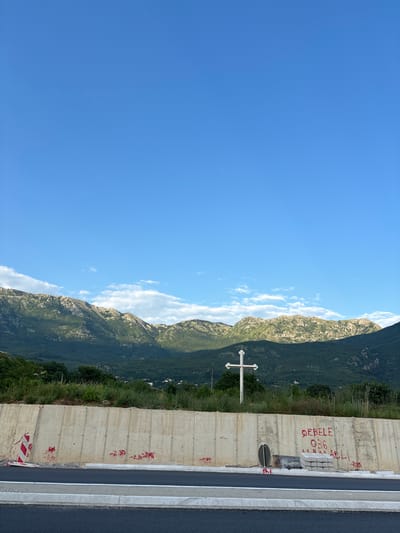 The image shows a wide shot of a rural landscape with mountains in the background and a concrete barrier in the foreground. The sky is a clear blue with some wispy clouds. A white cross stands on a grassy embankment behind the concrete barrier. The barrier itself is marked with red graffiti and some construction cones. The road in front of the barrier is paved and appears to be new. The scene suggests a location in or near Radanovići, Montenegro, likely along a roadway undergoing construction or maintenance.