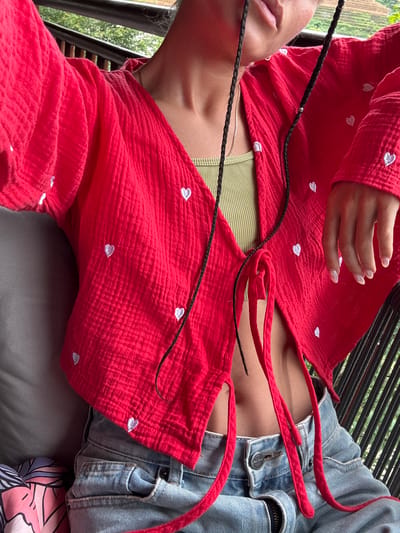 An individual is captured from the mid-torso upwards, seated or leaning back outdoors in Púng Luông, Vietnam. They wear an open-front, long-sleeved red shirt crafted from a textured, crinkled fabric, adorned with scattered small white embroidered hearts. Beneath it, an olive green ribbed crop top is visible, exposing a lean midriff. Light wash, distressed denim jeans are worn on the lower half, featuring a visible metallic button and zipper. The person's dark braided hair includes a strand with a small gold bead. A thin gold chain necklace is visible around the neck, and the lips are slightly parted. The right hand, with long, oval nails polished with a subtle iridescent or clear finish, rests gently on the red top. The background reveals lush green foliage and a dark metal railing with vertical slats, indicating an elevated outdoor setting. A gray upholstered surface and a colorful patterned fabric cushion are partially visible on the left side of the frame.