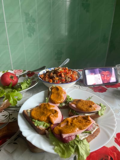 In an indoor setting, possibly a kitchen or dining area, a table is laid with a floral-patterned tablecloth. On the table, there are several open-faced sandwiches on a white plate. Each sandwich consists of a slice of dark bread topped with lettuce, a slice of pink ham or sausage, and a dollop of orange sauce. To the left of the sandwiches, a shallow bowl contains a mixture of chopped vegetables, likely a salad or stew. Adjacent to the vegetable bowl is a white plate with a single red tomato and lettuce leaves. In the background, a wall is covered with light green tiled squares. A mobile phone, placed on the table, displays a paused video or image of a person's face. The lighting suggests an indoor environment during the daytime. The scene does not provide specific visual cues to pinpoint the location within the city of Красное, Russia.
