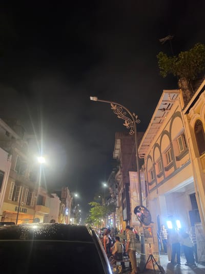The image shows a street scene at night in the city of Medan, Indonesia. The street is lined with buildings, some of which appear to be historic, with ornate architecture. There are streetlights illuminating the scene, and a bright light source is visible in the foreground, suggesting the presence of a photoshoot or filming activity. Several people are visible in the image, some standing and others seated. One group appears to be gathered around some equipment, possibly for a photo session. The road surface in the foreground is wet, indicating recent rain. Banners and signs are also visible on the buildings, some with Indonesian text. The overall atmosphere is one of a lively urban environment at night.