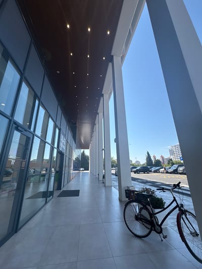This is a low-angle, eye-level shot of a modern building's exterior walkway with a view of a parking lot and city skyline in Bjeliši, Montenegro. The walkway is covered by a dark wooden ceiling with embedded lights and supported by large white pillars. To the left, a glass-fronted building with multiple doors and windows reflects the surroundings. A black bicycle with a basket on the rear is parked on the right side of the walkway. Beyond the walkway, a paved parking lot filled with cars leads to a street with a few buildings and trees visible in the distance. The sky is a clear, bright blue.