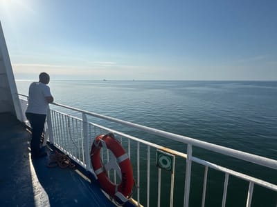A person stands on the deck of a ferry, looking out at the calm ocean. The sun is bright and high in the sky, casting long shadows on the deck. The sky is a clear, pale blue with a few wispy clouds. The ocean is a deep blue, with small ripples on the surface. In the distance, several ships can be seen on the horizon. A lifebuoy hangs on the railing, with "EMDEN" visible on it. A coiled rope lies on the deck near the railing. The overall atmosphere is peaceful and serene. The person appears to be contemplating the vastness of the sea.
