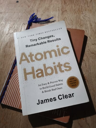 A white-covered book titled "Atomic Habits" by James Clear is positioned centrally, angled, atop another book with a reddish-brown cover, on a light brown wooden surface. The "Atomic Habits" cover displays "Tiny Changes, Remarkable Results" and "An Easy & Proven Way to Build Good Habits & Break Bad Ones," along with "#1 NEW YORK TIMES BESTSELLER" and an "OVER 1 MILLION COPIES SOLD" circular emblem. A bookmark with blue fringed tassels extends from between the two books, emerging from the top left corner. The wooden surface exhibits visible grain. Shadows are cast to the right and bottom right of the books, consistent with illumination from the left. The scene is located in General Trias, Philippines. No individuals are present.