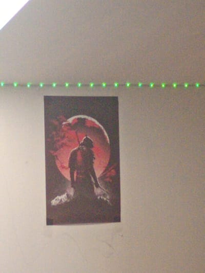 The media file shows a poster on a wall, illuminated by a string of green LED lights. The poster depicts a lone figure from behind, standing against a large, blood-red moon. The figure is silhouetted and appears to be wearing armor or a costume, with a weapon or staff held in one hand. The scene is dark and atmospheric, with the red moon casting a dramatic glow. The poster's style suggests a fantasy or science fiction theme. The LED lights create a vibrant, almost futuristic border around the top of the image, contrasting with the muted tones of the wall and the poster itself. The poster is hung on a light-colored wall, which appears to be indoors. The image is taken from a slightly upward angle. There is no visible text in the media file. The setting appears to be a room in Kobyłka, Poland.