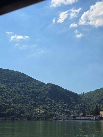 This image captures a scenic view of a lush, tree-covered mountain slope adjacent to a body of water. The mountain dominates the right side of the frame, its dense foliage creating a rich green texture that extends upwards towards a clear blue sky. A few scattered white clouds drift across the sky, suggesting fair weather.
Along the base of the mountain, and partially visible on its lower slopes, are buildings and structures that appear to be part of a small settlement or industrial area. These structures are nestled amongst the trees and along the water's edge. The body of water on the far right is a calm, greenish hue, reflecting the sky and surrounding greenery with subtle ripples on its surface. A man-made structure, possibly a dam or retaining wall with a series of visible supports, runs along the edge of the water and mountain.
The perspective suggests the photo was taken from a boat or from a similar vantage point on the water, looking towards the shore and the mountain. The lighting indicates it is daytime, likely late morning or afternoon, given the brightness of the sky and the illumination of the landscape. The scene evokes a sense of natural beauty combined with human presence in a waterside environment. There is no visible text in the image.