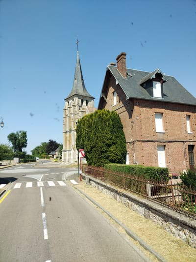 This is a photograph of a village scene in Saint-Aubin-d'Écrosville, France, featuring a historic church tower and a brick house. The church tower, constructed of stone, has a pointed roof and a clock face. Adjacent to the church is a large, dense green hedge. A red-brick house with white-shuttered windows and a dark grey tiled roof stands to the right of the church. A rusted metal fence borders the property. In the foreground, a paved road with white lane markings and a pedestrian crossing is visible. The sky is clear and blue. The overall environment suggests a sunny day in a rural French village.