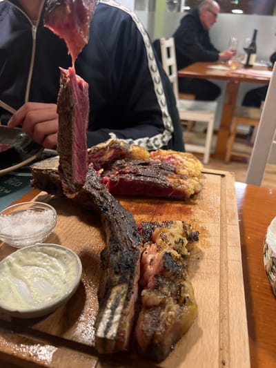 A close-up shot reveals a large, grilled steak on a wooden cutting board. The steak, cut into sections, displays a variety of textures, including crispy charred areas, succulent medium-rare portions, and visible fat. Accompaniments include small containers of salt and a creamy white sauce. A person holding a slice of steak is visible in the foreground, while in the blurred background, another person is seen at a table with wine glasses.