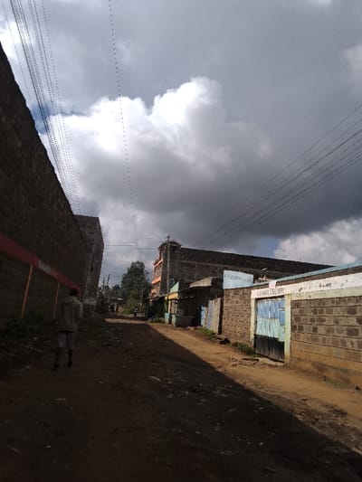 The image depicts a street scene in Njoro ward, Kenya, under a predominantly cloudy sky with patches of visible blue. A wide, unpaved dirt road, marked by tire tracks and foot traffic, slopes gently into the distance. A significant diagonal shadow covers the lower-right portion of the road.
In the immediate foreground, a male subject is observed from behind, walking away from the viewer towards the center of the frame. He wears a light-colored, long-sleeved shirt and dark shorts. Further down the road, additional indistinct human figures are visible.
To the left, a tall, unfinished brick wall with a distinct red horizontal stripe near its top edge extends along the road. Green vegetation is present at its base.
On the right side of the road, a shorter brick wall with a light-colored, possibly painted surface features a blue and black metal gate. White lettering above the gate reads "KAGWETI AUTO PARTS." Beyond this wall, a series of single-story commercial structures or shops with varying facades and door colors (including blue and green) line the road. Further back, a multi-story building with prominent orange and grey sections is visible. A tree with green foliage stands between buildings in the mid-ground.
Numerous black utility lines stretch across the upper portion of the frame, traversing the cloudy sky. The clouds range from bright white cumulus formations to darker, denser grey overcast areas, indicating variable weather conditions.
