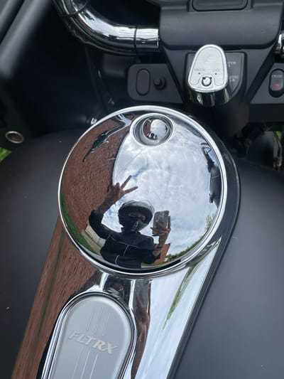 The media file shows a close-up, reflective view of a motorcycle's chrome gas cap. The highly polished surface of the gas cap acts as a mirror, reflecting the surrounding environment and a person taking the photograph.
**Subjects:**
* **Objects:** A chrome motorcycle gas cap with a circular shape and a small, centrally located button. The gas cap is surrounded by the black body of the motorcycle and chrome accents. To the left, a silver ignition switch assembly with "UNLOCK" and "LOCK" indicators is visible. To the right, a chrome panel with the embossed text "FLTRX" is present.
* **People:** A reflection of a person wearing a black motorcycle helmet and jacket is visible in the gas cap. The person is holding a smartphone and making a peace sign with their right hand.
* **Scene/Setting:** The reflection shows a brick building with windows and a cloudy sky. Green grass and possibly trees are also reflected, suggesting an outdoor setting.
**Activity/Event:**
The image captures a moment where the photographer is taking a selfie using the reflective surface of their motorcycle's gas cap.
**Notable Details:**
* **Time of Day/Weather:** The cloudy sky suggests it is daytime.
* **Location Cues:** The presence of a motorcycle and the brick building are the primary environmental cues. The specific location within Oktyabr'skii, Russia, is not visually discernible from the image itself, but is provided as context.
* **Emotions:** The peace sign suggests a casual and positive mood.
**Visible Text:**
* "UNLOCK"
* "LOCK"
* "FLTRX"
* "CRUISE" (on a button above the ignition)
