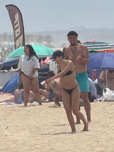 The image depicts a crowded beach scene at Espinho, Portugal, under a bright sky. In the foreground, a woman in a black bikini walks on the sand, her body angled towards the right side of the frame. Her dark hair is pulled back, and she is wearing a dark baseball cap. Behind her and to her right, a shirtless man with dark hair and a beard smiles and claps his hands, wearing turquoise swim trunks. To the left of the woman in the bikini, a woman in a white t-shirt and black bikini bottoms stands looking to the left. A large, dark flag with white lettering is positioned to the left of the women. Beach umbrellas, some colorful and some blue, are visible in the background, along with other beachgoers and the ocean. The sand is a light brown color.