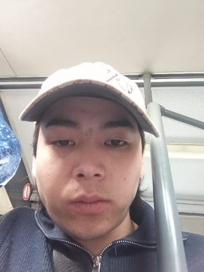 A close-up, low-angle self-portrait depicts an individual, an adult male, likely of East Asian or Central Asian ethnicity, in an indoor environment resembling public transport. He is positioned centrally, wearing a light-colored baseball cap with a dark brim and a dark blue ribbed zippered jacket. His eyes are directed downwards, and he maintains a neutral facial expression with visible stubble on his chin and jawline. To the right of the individual, a vertical gray metallic pole extends. To the left, a partial view of a blue object with a swirling pattern and a circular red and white sign are visible. The ceiling above is light-colored. The image does not contain discernible visual information to confirm the city location of Urtaaul, Uzbekistan.