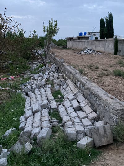 The image depicts an outdoor scene in Qila Saifullah, Pakistan, characterized by a mix of natural elements and human construction materials. In the foreground and mid-ground, a large quantity of light grey concrete blocks are scattered and piled unevenly on the ground, some lying flat, others tumbled. These blocks appear to have either fallen from an adjacent structure or are awaiting use in construction. Green grass and weeds grow amongst the blocks and on the lower left side of the frame.

To the right of the scattered blocks, a low, unfinished concrete retaining wall runs parallel to a dirt path. The path itself is dry and dusty, with sparse patches of green vegetation. Further along the path and in the background, several trees with green foliage are visible, along with some barren or dead tree branches on the left.

In the mid-ground and background, there are several buildings, likely houses or other structures. One prominent building has a flat roof with at least two blue water tanks on top, a common feature in the region. Another building features a flat roof and small rectangular openings, possibly windows or vents. A concrete wall separates some of these structures from the dirt path. Tall, slender evergreen trees (resembling cypress trees) stand behind or next to these buildings. The sky is overcast with soft, diffuse light, suggesting either an early morning, late afternoon, or generally cloudy day. No people or visible text are present in the image. The scene conveys an impression of an undeveloped or semi-developed area, possibly undergoing construction or experiencing neglect.