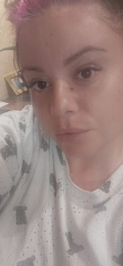 The image shows a close-up, selfie-style photograph of a woman's face. She has light skin and is wearing a white top with a gray teddy bear pattern. Her hair is styled up, with some pink-colored strands visible at the top of her head. She has dark eyebrows and is wearing what appear to be false eyelashes. The background is blurry and appears to be an indoor setting, possibly a home. A small framed picture is visible on the left side of the image, in the background. The lighting suggests it is daytime, but the source of light is not clear. The photograph was taken in Marynivka, Ukraine.
