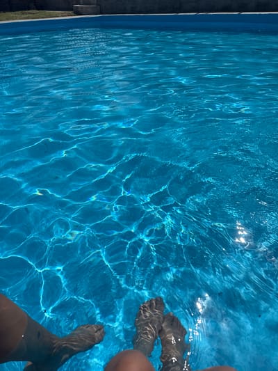 The image shows two pairs of bare feet submerged in the clear blue water of a swimming pool. The water surface is rippled, creating interesting light refractions. In the background, the edge of the pool and a grassy area can be seen. The perspective is from someone sitting at the edge of the pool, with their legs extended into the water. The location is identified as San Baltazar Torija, Mexico.