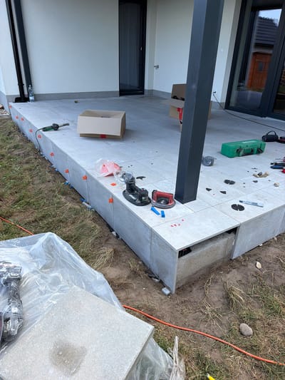 The image depicts an outdoor construction scene in Dolní Vinice, Czechia, focusing on the installation of large, light gray tiles on an elevated patio or building facade. The tiles are being laid with precision, evidenced by numerous red and blue tile spacers visible between them. A black metal support pillar stands prominently in the foreground, integrated into the tiled structure. Construction tools and materials are scattered around the area, including a green toolbox, a heat gun, a power grinder with a circular blade, a roll of black tape, a plastic bag containing orange cones, and various smaller construction aids. Several cardboard boxes, one of which is open, suggest recent deliveries or ongoing work. The surrounding ground is a mix of dry grass, soil, and some small stones, indicating an unfinished or recently excavated area. The background shows a portion of a modern building with large windows, and in the reflection of the glass, a small wooden shed can be seen. The overall scene suggests a construction or renovation project in progress.