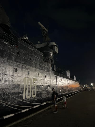 The image captures a nighttime view of a large, dark-hulled ship docked at a pier. The ship has the numeral "105" painted in white on its side. Two people are visible on the pier: an adult male wearing a dark shirt and shorts, and a child riding a scooter, wearing a white shirt and red shorts. The pier is lit by artificial lights, and other ships and structures are visible in the background. The overall scene suggests a maritime location, possibly a museum or a working port at night. The location is identified as Verkhnyaya Stavropol'ka, Russia.