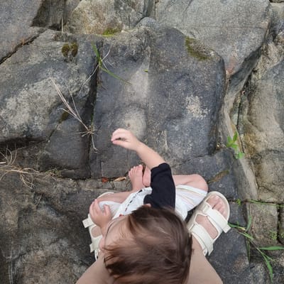 This image depicts a child sitting on a large, textured rock formation. The child is seen from above, with their head tilted and their right hand reaching out to interact with something small and green, possibly a leaf or a sprout, which is not clearly identifiable. The child is wearing a black long-sleeved shirt and striped shorts, and their feet are bare, adorned with white sandals. The background consists of dark, grey rocks with patches of moss and sparse dry grass growing in crevices. The environment appears to be natural, possibly a park or outdoor recreational area with rock formations. The image does not contain any elements that would place it specifically within Mureungdowon-myeon, South Korea.