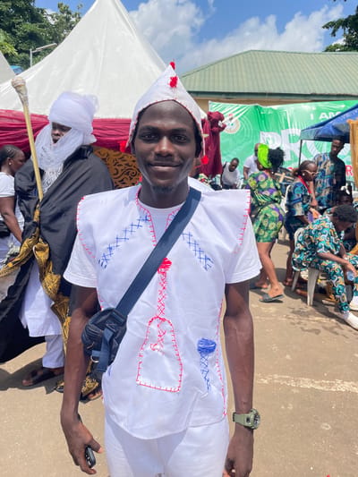 A young Black man in a white outfit with red and blue stitching stands in the foreground, smiling at the camera. He wears a white conical hat adorned with red tassels. A blue crossbody bag is slung over his shoulder. Behind him, a taller Black man in traditional white attire, including a turban and a long black robe with gold trim, holds a staff with a decorative top. Several other people in colorful clothing are visible in the background, some standing and some seated. A large green banner with text is displayed behind them. The scene appears to be outdoors, possibly at a festival or gathering, under a bright sky with some clouds. The ground is paved, and there are tents and structures suggesting an event space. The overall setting suggests an outdoor cultural event in Alagba, Nigeria.
