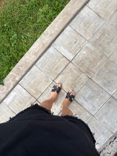 The image is a top-down, full-body shot of a person standing on a paved walkway in Verkhneyarkeyevo, Russia. The person is wearing black sandals and a black, frayed-hem skirt. The walkway is made of light-colored rectangular tiles arranged in a herringbone pattern. To the left of the walkway is a strip of green grass with some sparse weeds. The scene appears to be outdoors, possibly in a park or residential area.