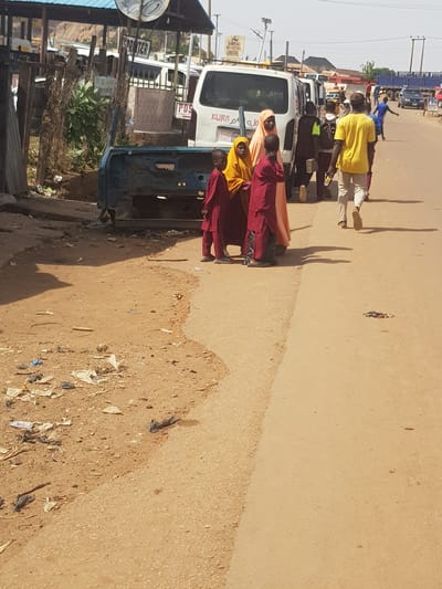 This image captures a vibrant street scene in Kufena, Nigeria, under bright sunlight, indicated by the long, distinct shadows suggesting either early morning or late afternoon. The setting is an unpaved road with sections of asphalt, flanked by buildings and sparse vegetation.
In the foreground, a group of three children or young people stands near a white van, two wearing red robes and one in an orange-yellow robe and head covering, possibly indicating religious attire or school uniforms. Next to them is a partially disassembled blue vehicle, resembling an old car chassis. Litter, including plastic bags and debris, is scattered along the dirt shoulder of the road.
Behind the robed individuals, several people are visible. A man in a bright yellow shirt and light-colored pants walks in the mid-ground. Further back, more individuals are seen, some near parked vehicles. A person in a blue shirt is in the distance. The street is active with various vehicles, including several vans and what appears to be a blue flatbed truck in the background.
Visible text includes "KURDA" and "POS" on the white van. A sign for "KASHAWA SOVENTURES LIMITED" is legible, along with another sign that reads "SPRITER". Utility poles and power lines crisscross the upper part of the scene. The overall atmosphere depicts a bustling street in an urban or semi-urban Nigerian environment.