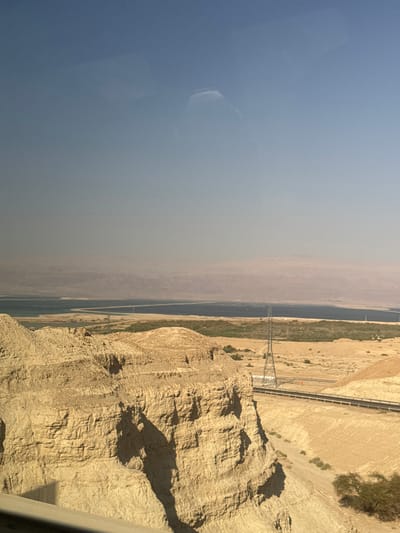 This image captures an aerial or elevated view of a desert landscape with a prominent body of water. The scene is dominated by dry, sandy terrain and rugged, rocky cliffs. A long, narrow body of water, likely a river or canal, runs through the arid environment, flanked by patches of green vegetation. A man-made structure, resembling a bridge or elevated roadway, crosses over the water. In the distance, a hazy sky suggests a warm, possibly dusty atmosphere, typical of a desert environment during daylight hours.
The composition of the image suggests it was taken from a moving vehicle or a high vantage point. The sunlight casts distinct shadows on the rocky surfaces, indicating it is daytime. The muted colors and the vastness of the landscape evoke a sense of remoteness and the harsh beauty of the desert. The presence of the water body and the infrastructure signifies human presence and activity in this otherwise desolate setting. The overall impression is one of a landscape shaped by both natural forces and human engineering.