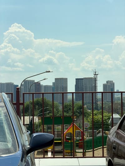 The image shows a view from a high vantage point looking out over a city. In the foreground, a red metal fence partially obstructs the view. To the left, the side of a dark blue car is visible. To the right, the side of a dark brown car is visible.
Beyond the fence, a playground with colorful structures, including a yellow and red playhouse and green fencing, is visible. A basketball hoop is also present. Behind the playground, dense green trees line the landscape.
In the mid-ground, several tall, modern apartment buildings dominate the skyline. These buildings have a uniform appearance with many windows. A tall, dark metal tower with antennas and equipment is positioned among the buildings. Streetlights with curved poles are also visible in front of the apartment buildings.
The background consists of a vast expanse of sky filled with large, fluffy white clouds against a pale blue backdrop. The overall impression is an urban scene on a bright, partly cloudy day. The location is identified as the city of Azov, Russia.