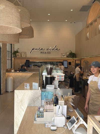 The image depicts the interior of a brightly lit coffee shop. In the foreground, a counter is made of light-colored, veined marble and wood. On the counter, there are various items, including a small scanner, a tablet computer, a white printer, and packets of coffee or tea. To the right, a young woman wearing an apron with the name "BETH" visible, and a baseball cap, is looking towards the right and smiling. Behind her, further into the shop, are other employees working at a coffee bar. A professional espresso machine is visible on the left side of the counter, with a grinder filled with coffee beans. The background features a wooden wall with the words "pura vida MIAMI" in script lettering. The ceiling has large, woven pendant lights. To the far left, a white trash receptacle is visible near a stone wall. The scene suggests a bustling cafe environment.