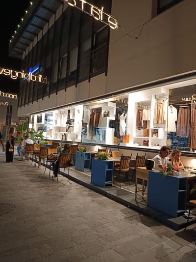 The image depicts an outdoor dining area in front of a boutique store at night. The store's facade is illuminated, showcasing mannequins and clothing displays. Several tables are set up on the cobblestone sidewalk, with patrons seated and dining. In the foreground, a couple is seated at a table, with the man looking at his phone and the woman looking away. In the middle ground, other patrons are also seated at tables. A woman in a black dress is standing to the left of the frame, and a man is seated at a table to her right. The scene appears to be in the city of Roseto degli Abruzzi, Italy, given the context provided by the prompt, suggesting a lively evening atmosphere in a commercial area.