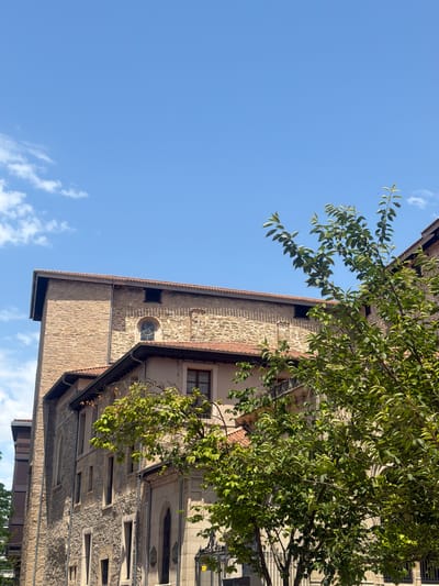 The image shows a stone building with a red-tiled roof, seen from a low angle against a clear blue sky with a few wispy clouds. A tree with lush green leaves hangs into the frame from the top right, partially obscuring the building. The building's facade is constructed from rough-hewn stones and features multiple windows of varying sizes, some with decorative stone surrounds. One small, round window with a glass pane is visible on the upper section of the building. The sunlight casts shadows on the stone, indicating it is daytime. The overall impression is of an old, possibly historic, building in a sunny, outdoor setting. The location context is Vitoria-Gasteiz, Spain.