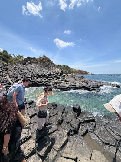 The image depicts a group of people at a seaside location with unique geological formations. The scene appears to be a rocky coastline with hexagonal basalt columns, characteristic of places like Gành Đá Dĩa in Phú Lương, Vietnam.
Several individuals are visible, some are sitting and others are standing. One woman, wearing a sun hat and a patterned dress, is looking out towards the sea. A man in a blue shirt is also present, and other people are partially visible, suggesting a gathering or a shared outing. One person is wearing a white hat. There's a straw bag resting near some of the individuals.
The sea is a vibrant turquoise color, with waves gently breaking against the dark, geometric rocks. The sky is a clear blue with scattered white clouds, indicating a sunny day. The sun appears to be high in the sky, suggesting it's daytime, likely midday or early afternoon. The atmosphere seems relaxed and leisurely, with people enjoying the natural scenery. There is no visible text in the image.