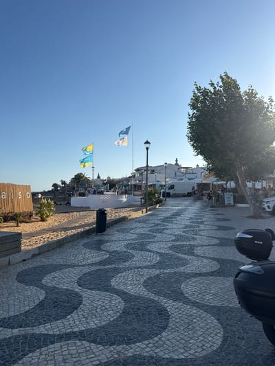 This is a vertical, eye-level shot of a waterfront promenade in Lagos, Portugal, on a sunny day. The scene depicts a paved walkway with a distinctive wave-like pattern created by dark and light grey cobblestones.

On the left side of the promenade, a clear blue sky dominates the upper portion of the frame, with the upper branches and leaves of a large tree casting dappled sunlight. Beneath the tree, buildings with white facades and awnings line the promenade, with some people seated at outdoor tables, suggesting a cafe or restaurant setting. A white van is parked along the sidewalk.

Further down the promenade, several flags are flying from poles. Two flags on the left are white with blue emblems. Two flags on the right are yellow with blue wave-like designs. Beyond the flags, more buildings and palm trees are visible, hinting at a coastal town. The ground transitions from the cobblestone promenade to a sandy area with wooden planks and vegetation, including palm saplings. A small wooden structure with bamboo-like siding is visible in the foreground, bearing the partially visible text "aiso".

In the upper right corner of the image, the top of a black scooter or motorcycle is visible, with the text "H29" on its side. A dark green trash can is also present on the promenade. The overall atmosphere suggests a relaxed, daytime scene in a popular tourist area. The bright sunlight and clear sky indicate fair weather.