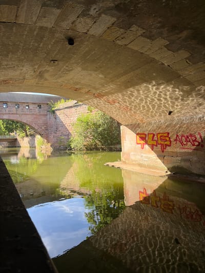 The image shows a view from underneath a stone bridge looking out towards a body of water, likely a canal or river. The bridge has a rough-hewn stone arch construction, with sunlight dappling through the arches onto the underside of the bridge and the wall of the bridge abutment. The wall of the abutment on the right side of the image features graffiti in red and yellow paint. The water is a murky green color and reflects the bridge, surrounding foliage, and sky. In the distance, another stone bridge and a building with a tiled roof can be seen, suggesting an urban environment. This scene is located in Nuremberg, Germany, along the Pegnitz river where it flows beneath the Henkersteg bridge. The Henkersteg is a historic footbridge in Nuremberg, Germany, that crosses the Pegnitz river. It connects the old town to the island of Schütt. The bridge is made of stone and is covered by a roof. The graffiti on the bridge abutment reads "444 ACAB!".