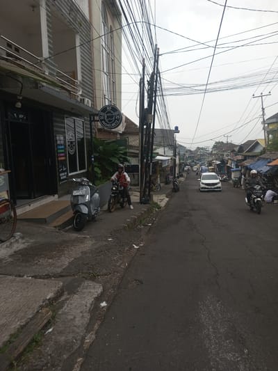 The image depicts a street scene in the city of Houaythat, Laos, under an overcast sky. The foreground shows a concrete sidewalk adjacent to a narrow, dark asphalt road with visible cracks. On the left, a dark-colored building with large glass windows and a prominent round sign advertising "PROHELMETZ" is visible. A light grey scooter is parked on the sidewalk in front of this building. Further along the sidewalk, a person wearing a red and white jacket and a helmet is seated on a dark motorcycle with gold-colored wheels. Numerous tangled utility wires span across the sky from multiple poles.
In the midground, a white car drives towards the viewer on the paved road, with multiple motorcycles and other vehicles visible further down the street. The right side of the street features a row of structures, some appearing to be makeshift stalls with tarpaulin roofs. Another individual, wearing a white helmet and dark jacket, rides a white and black motorcycle away from the viewer on the right side of the road. The background reveals a dense cluster of buildings, characteristic of an urban or semi-urban environment, extending into the hazy distance.