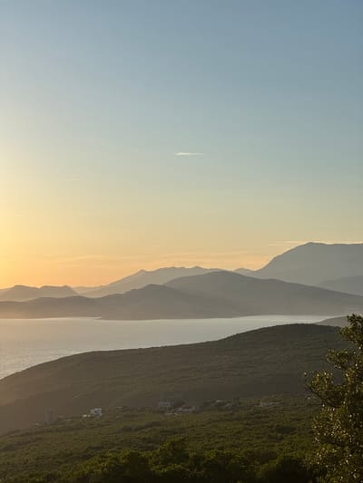 This is a vertical, wide-angle, eye-level, full shot of a serene coastal landscape at dusk in Kovačko Polje, Montenegro. The scene is dominated by layers of silhouetted mountains receding into the distance, their peaks softened by the fading light. A calm body of water, likely the Adriatic Sea, occupies the middle ground, reflecting the soft hues of the sky. The foreground is a lush, green hillside covered in dense vegetation, with a few scattered white buildings nestled amongst the trees, suggesting a small settlement. The sky transitions from a pale, soft yellow near the horizon, where the sun has recently set, to a clear, light blue overhead. A faint wisp of a cloud and two faint contrails from aircraft are visible in the upper sky. The overall mood is peaceful and tranquil, characterized by muted colors and soft light.
