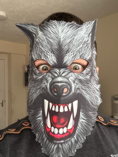 A person is wearing a werewolf mask. The image captures the person's eyes through the eyeholes of the mask. The mask features a grey and white werewolf face with a menacing open mouth revealing teeth and a red tongue. The surrounding environment includes a door, a wall and an item of clothing, which appears to be a black knitted garment with button detailing. The image does not provide enough information to identify the location as Welshpool, United Kingdom.
