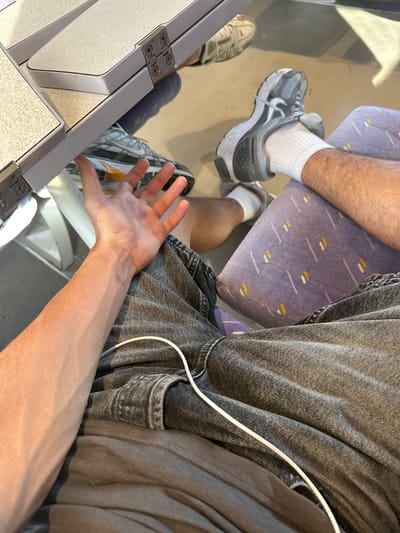 The image displays a close-up view of a person's forearm and hand resting on a folded-down tray table. The hand is open, palm facing upwards, with fingers slightly curled. The forearm shows visible veins and a light tan complexion. The person is wearing dark-wash denim jeans, and a white cable is draped across the jeans. In the background, a person's legs are visible, wearing athletic shoes and white socks, seated on a purple patterned seat. Another pair of athletic shoes is visible further back. The tray table is made of a light-colored material with metal hinges. The environment appears to be indoors, possibly on public transportation or in a waiting area, with a beige floor. The image's origin in Rivesaltes, France, is not discernible from the visual content itself.