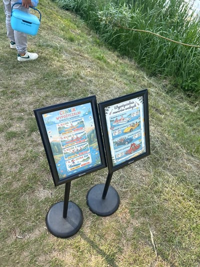 The image shows two framed signs on stands outdoors, likely in the city of Kobyla Góra, Poland, near a body of water, as indicated by the context of the signs which detail rental prices for water sports equipment. The sign on the left is titled "CENNIK WYPOŻYCZALNI SPRZĘTU WODNEGO" (Price list for water equipment rental) and lists prices for kayaks and a SUP board. The sign on the right is titled "Wypożyczalnia rowerów wodnych" (Water bike rental) and displays prices for different types of water bikes. In the background, there is grassy terrain and lush green vegetation. In the foreground, a person is partially visible on the left, holding a light blue cooler bag labeled "Arctic Breeze." The overall scene suggests a rental service for recreational water activities.