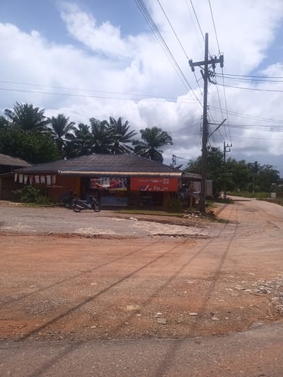 The image captures a rural scene in Plai Phraya, Thailand. A small, single-story building with a dark tiled roof sits on the corner of a dirt road. The building has a faded orange facade and a large banner with Thai writing and images covering the front. Two motorcycles are parked near the building's entrance. Laundry hangs on a line along the side of the building.

A weathered wooden utility pole stands next to the building, with numerous wires extending from it. The road is unpaved and reddish-brown, with visible patches of gravel and dirt. A denser cluster of trees, including palm trees, is visible behind the building. The sky is partially cloudy, with patches of blue visible between the clouds. The overall impression is of a quiet, rural environment with modest infrastructure.